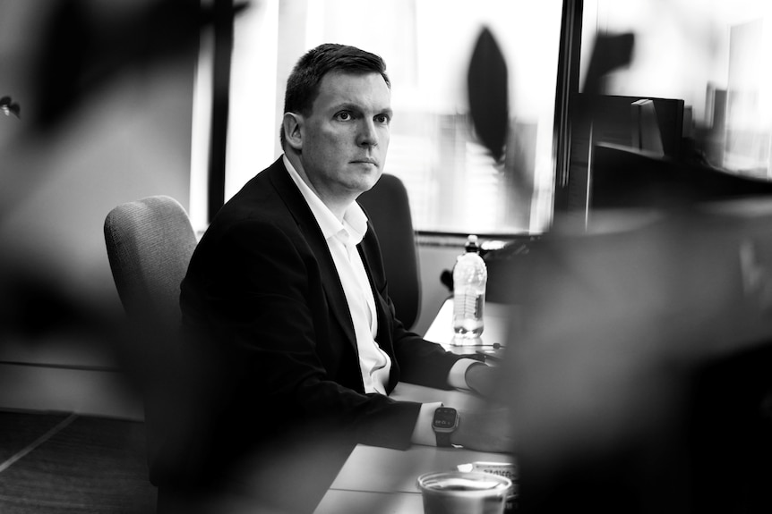 A black-and-white shot of a dark-haired man in a dark suit sitting at a desk in an office.