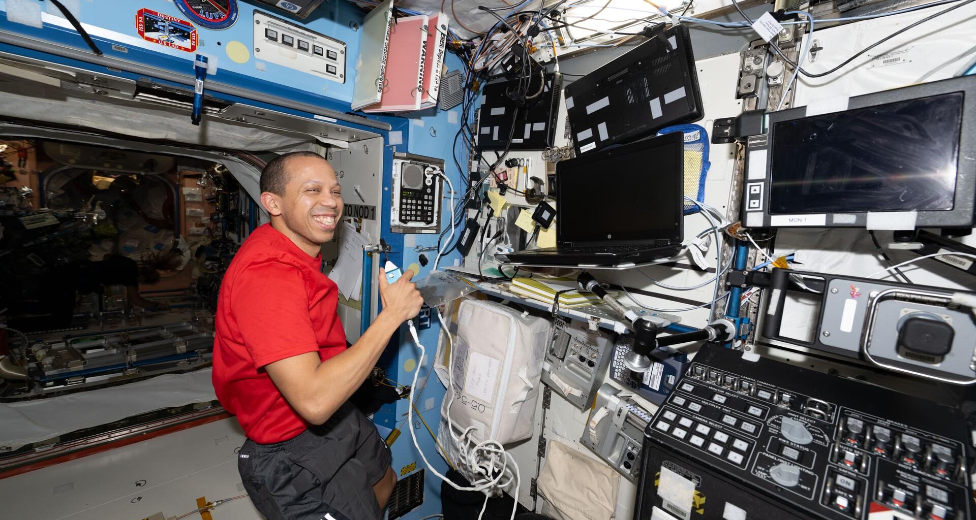 NASA astronaut Chris Williams is seated inside the Destiny laboratory module aboard the International Space Station. He is wearing a red shirt and black shorts with three white stripes underneath the right pocket. Chris is holding a device with a cord in his left hand and there are blank computer screens in front of him. He is smiling brightly and looking directly at the camera.