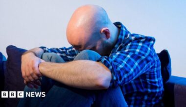 A man is hunched over on a sofa with his head between his knees and his arms locked around them.