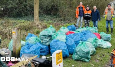 Three people, standing by a large amount of rubbish, including blue and green bin bags.