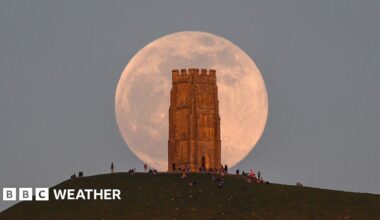 A huge pink hued Moon sits in a grey sky, behind the tower on Glastonbury Tor with people milling around at the base