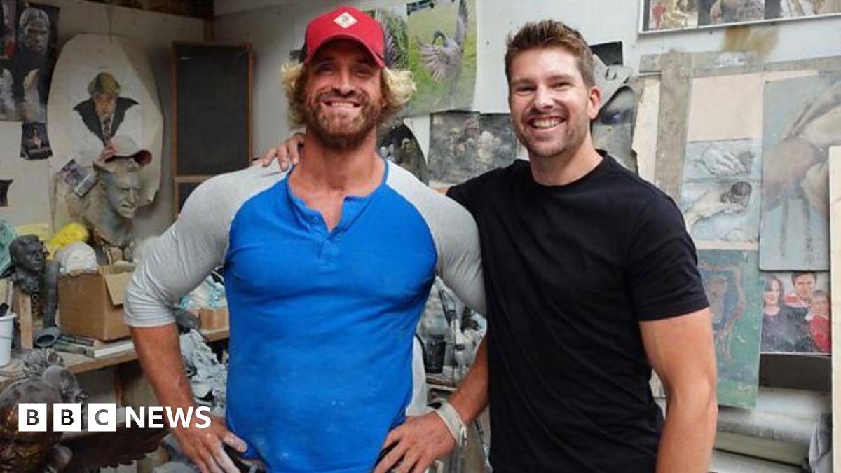 Nick Elphick wears a blue and grey long-sleeved top and red baseball hat. He is standing next to Darryn Frost, who has his arm around him. He is wearing a black T-shirt. They are both smiling at the camera and standing in an art studio, with paintings and small sculptures in the background.