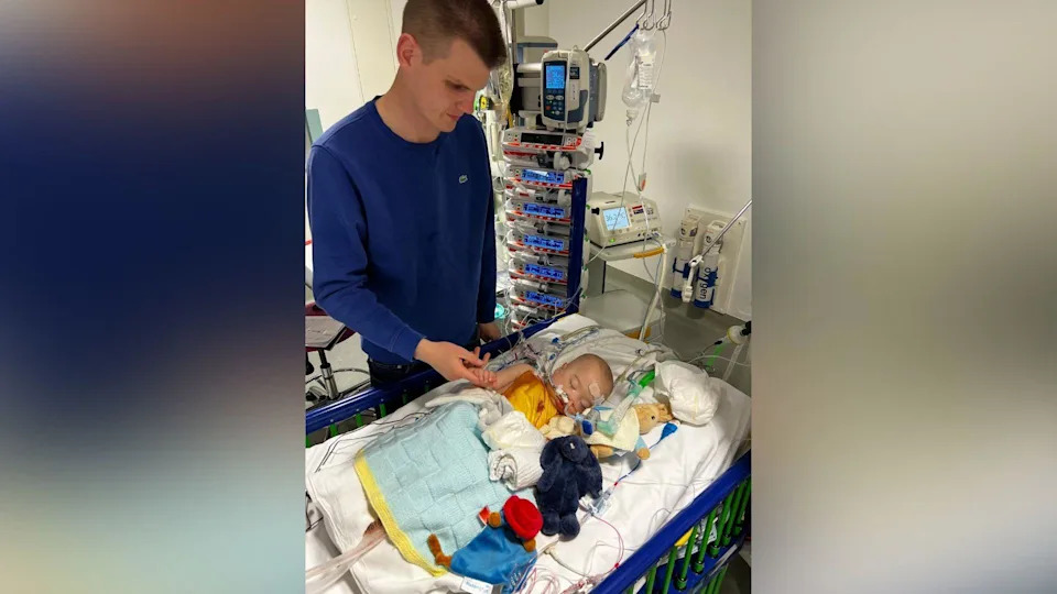 Alaric as a baby in a cot, surrounded by cuddly toys. He is in a hospital ward, and has tubes coming out of his nose. His father holds his hand and the room is full of medical monitoring equipment.