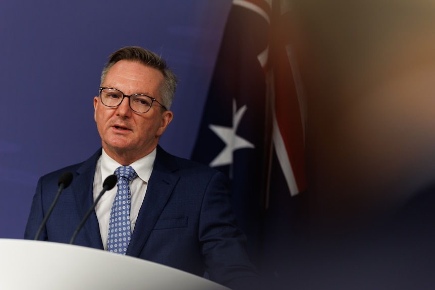 Chris Bowen looks slightly fed up as he stands in front of an Australian flag at a press conference.