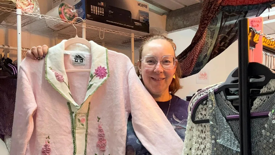 Rachel Grady smiles directly at the camera. She wears pink square glasses and smile at the camera. She holds up a pink and white cardigan in her left hand and stands behind a clothes rail at the forefront on the right. 