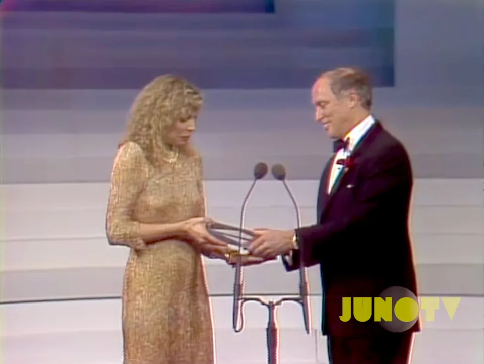 Two people on stage at an award ceremony, one in a formal dress and one in a tuxedo, holding a trophy