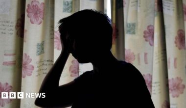 A silhouette of a woman holding her hand against her head. She is standing in front of floral curtains which are drawn.