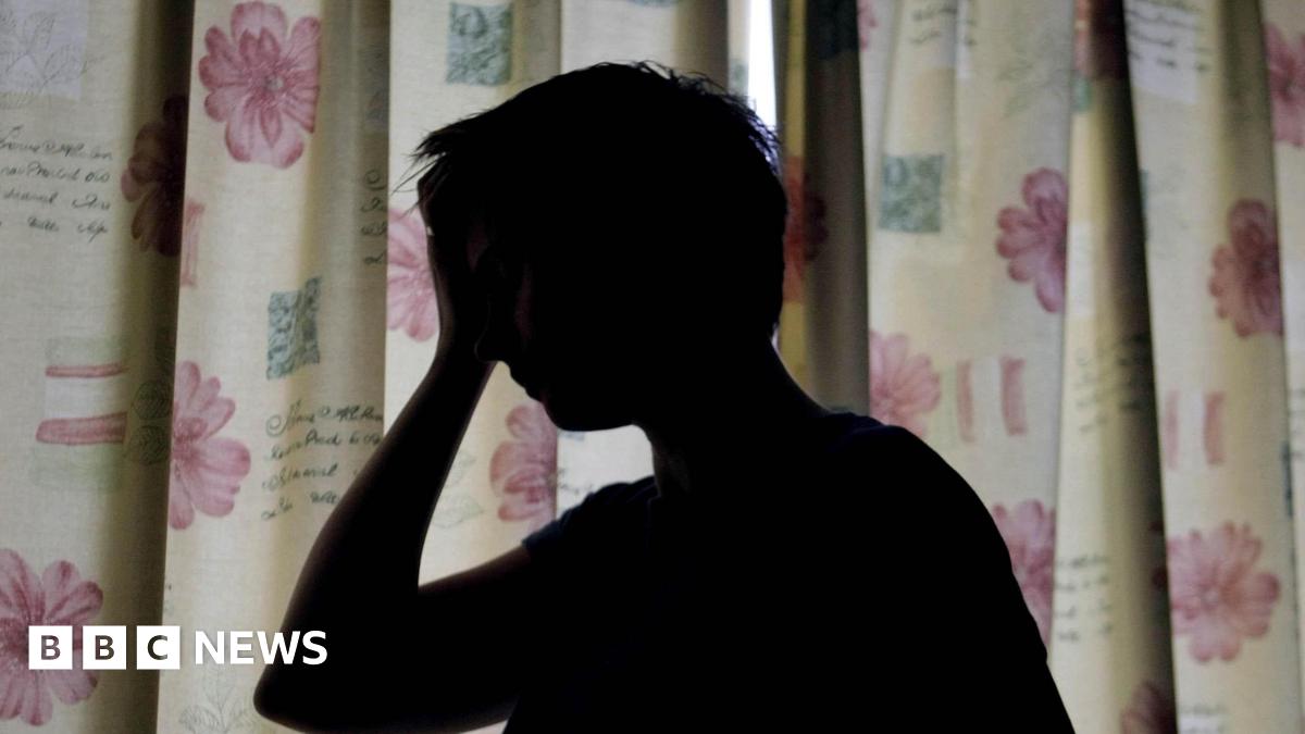 A silhouette of a woman holding her hand against her head. She is standing in front of floral curtains which are drawn.