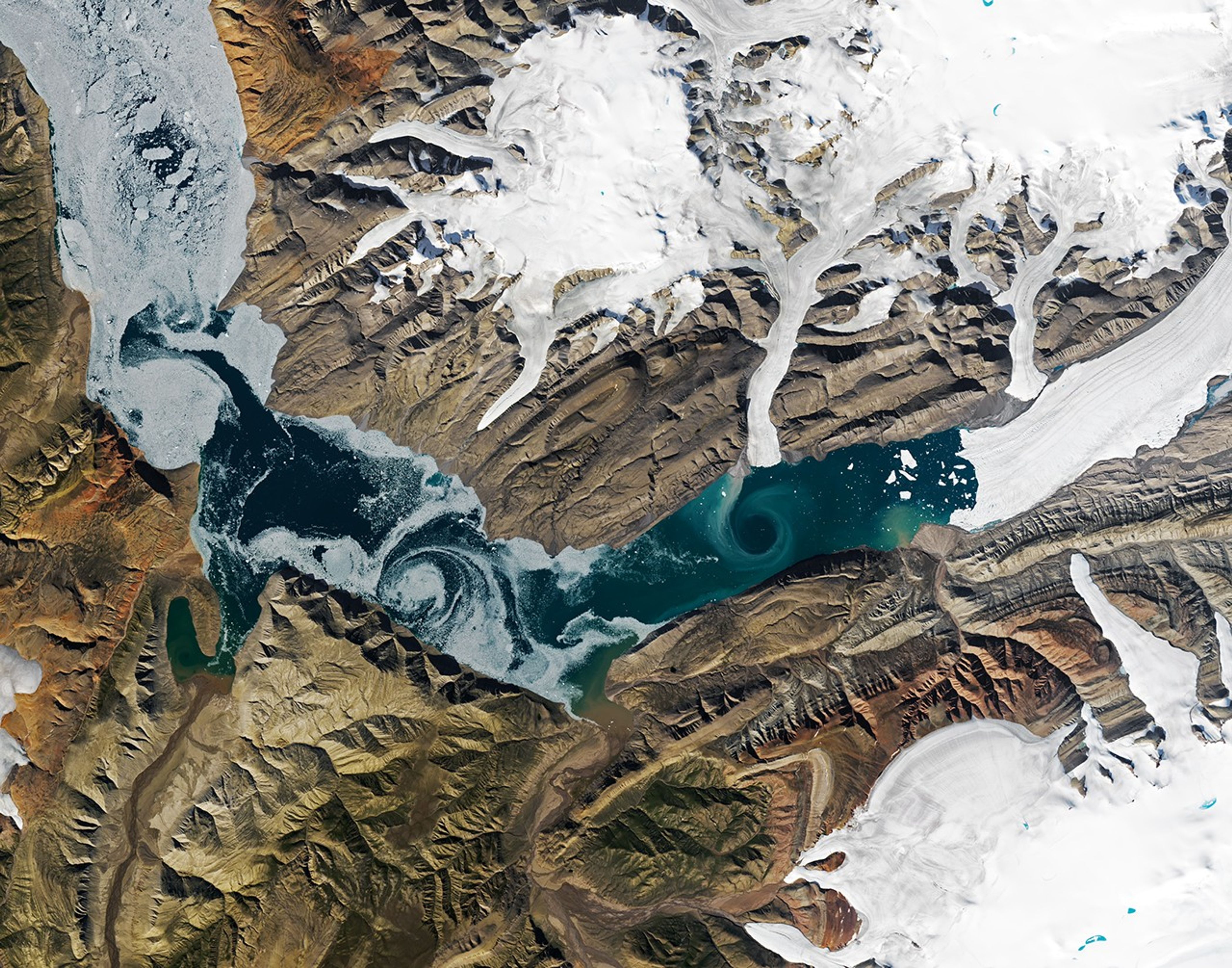 A V-shaped fjord cuts through barren brown land, with one patch of swirling water marked by white sea ice and another one colored turquoise by suspended sediment. Glacial ice flows into the fjord in several places.