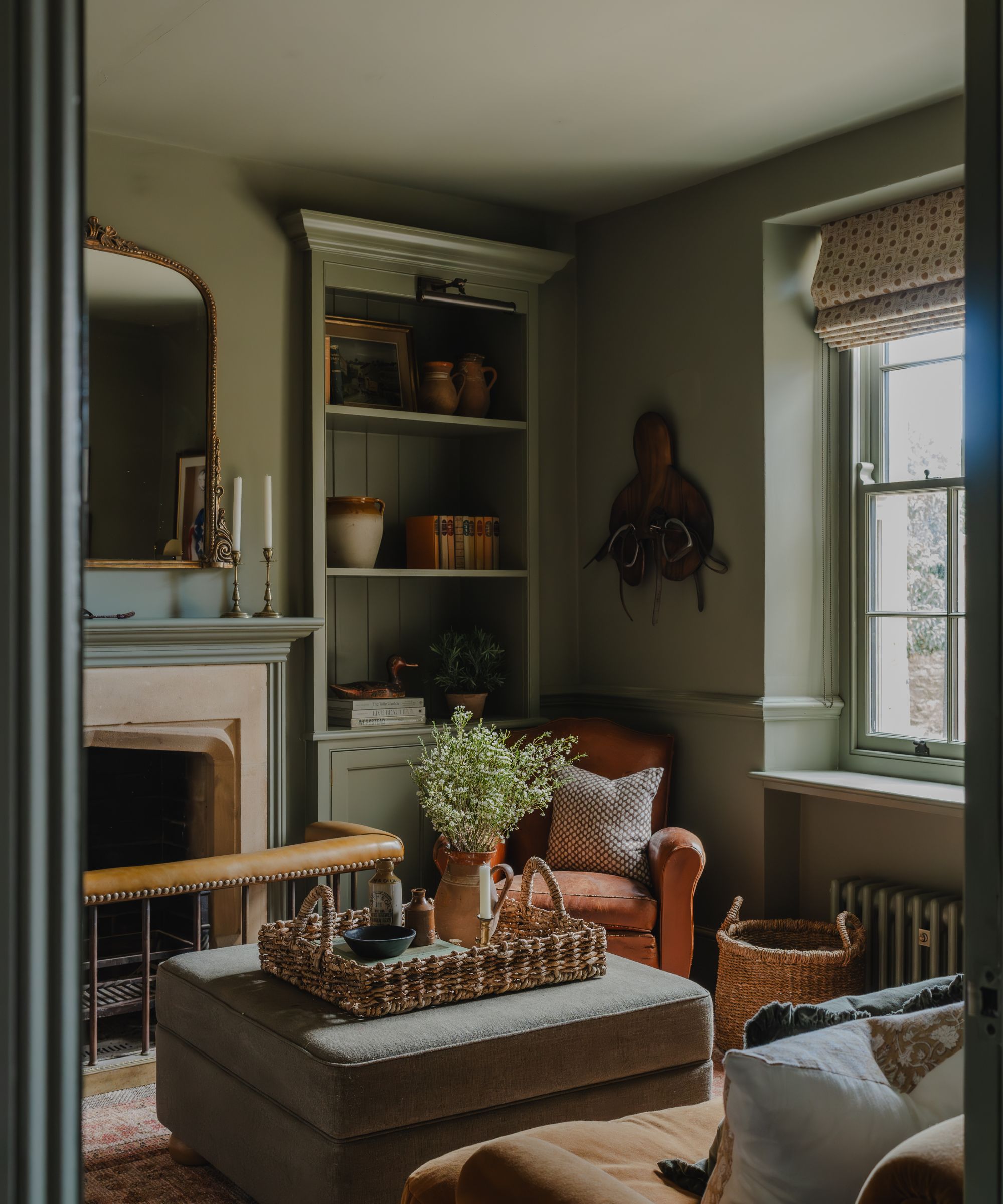 Living room with grey-green painted walls, a fireplace with a seat, an ottoman with a rattan tray filled with decor, a leather armchair in the corner, and a built-in bookcase with pots and books