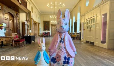 A small Peter Rabbit sculpture, positioned alongside a larger Peter Rabbit sculpture. They are in the centre of a large room in Worcester's Guildhall. The small sculpture is wearing a blue coat, the larger one is wearing a pink coat and is decorated with a floral pattern.