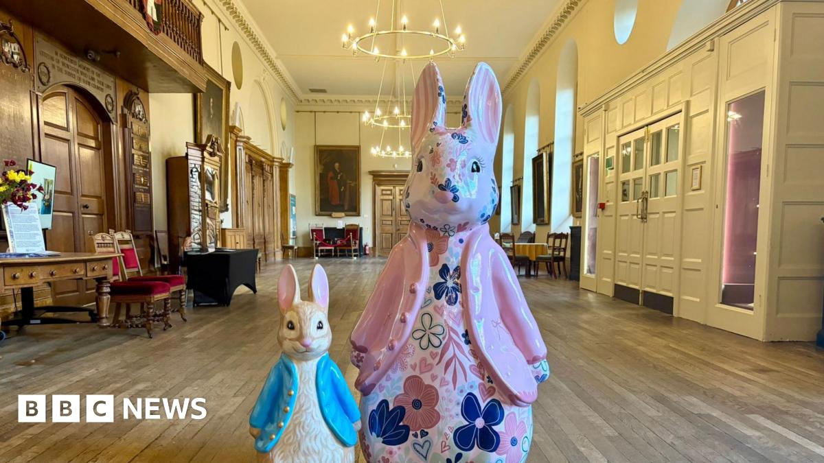 A small Peter Rabbit sculpture, positioned alongside a larger Peter Rabbit sculpture. They are in the centre of a large room in Worcester's Guildhall. The small sculpture is wearing a blue coat, the larger one is wearing a pink coat and is decorated with a floral pattern.