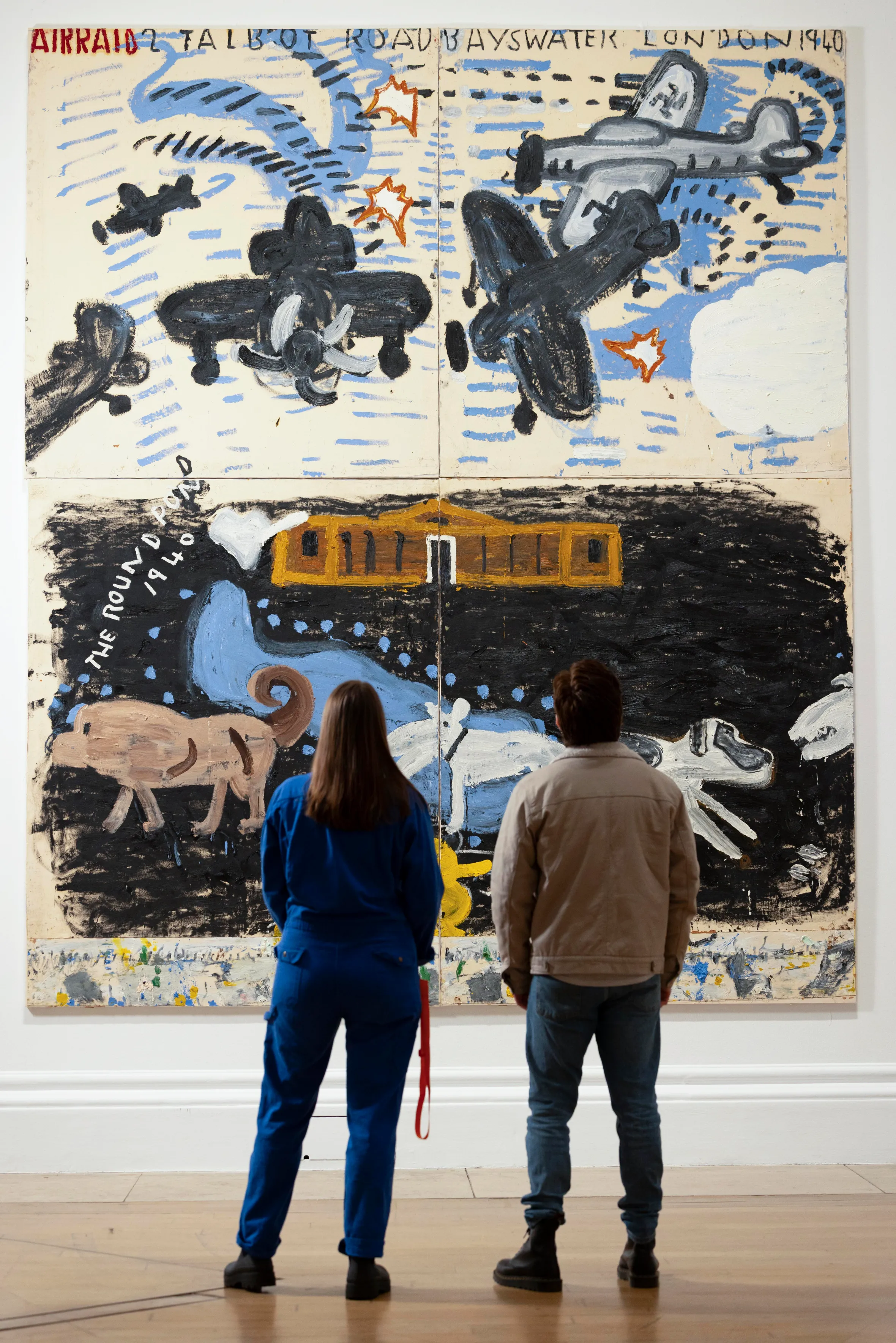 Two people view Rose Wylie's painting "The Picture Comes First," which depicts airplanes, a building, and dogs.