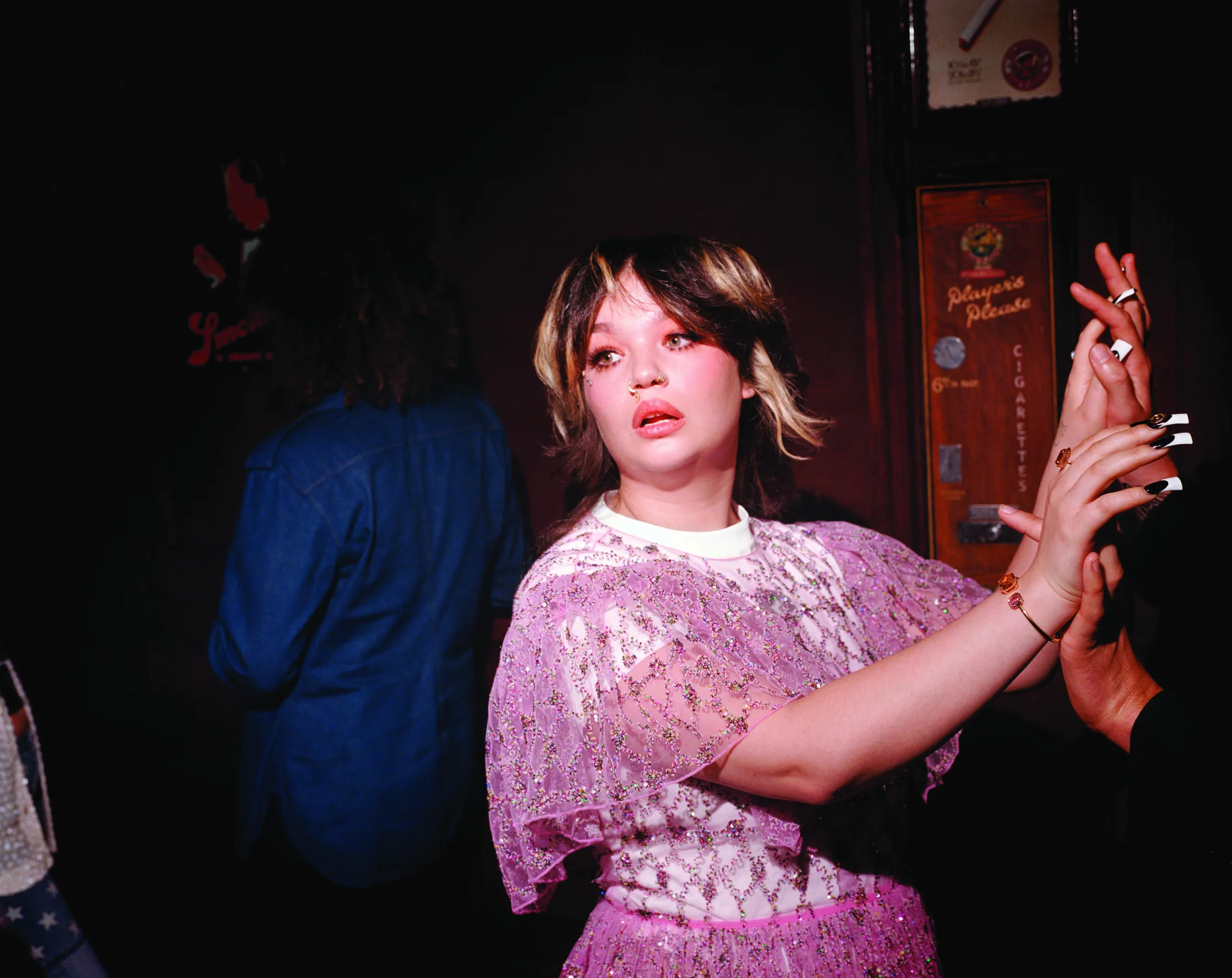 Lola Young in a sequined pink dress, gold nose rings, and bold eye makeup, holding another person's hands.