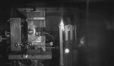 A close-up, black and white image of a scientific experiment setup, showing a bright light source in the center, possibly a flame or spark, surrounded by various metallic components and wires.