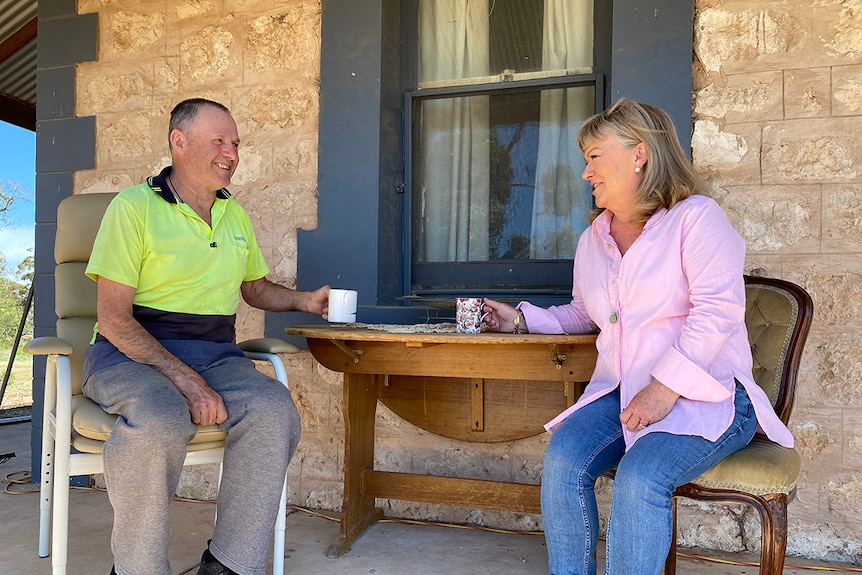 Man and woman sit at a table on the verandah of an old stone house drinking a cuppa.