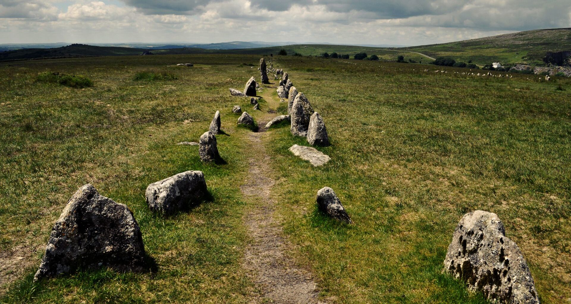 A view of lush rolling hills under a cloudy gray sky. In the foreground is a faded narrow dirt road with stones on either side of it