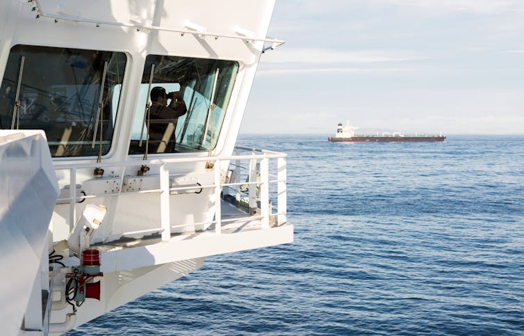 a man inside the bridge of a large ship at sea looks through binoculars with another ship in the background