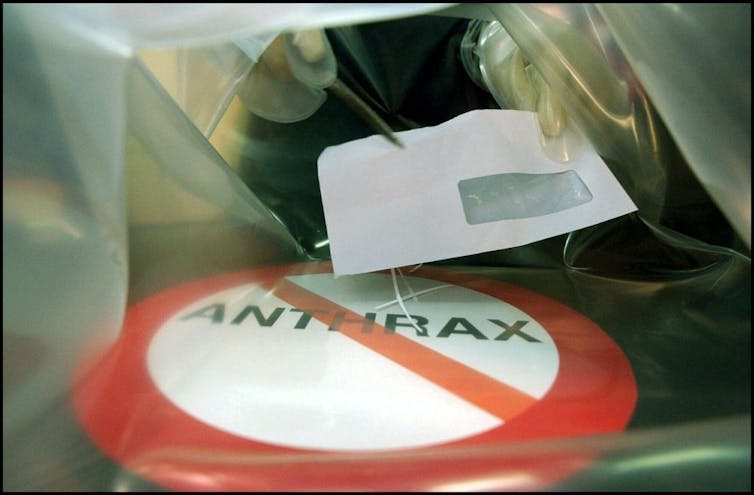 Close-up of person handling envelope with gloved hand and pliers held in a plastic cover over a platform with the word 'ANTHRAX' in a red no symbol