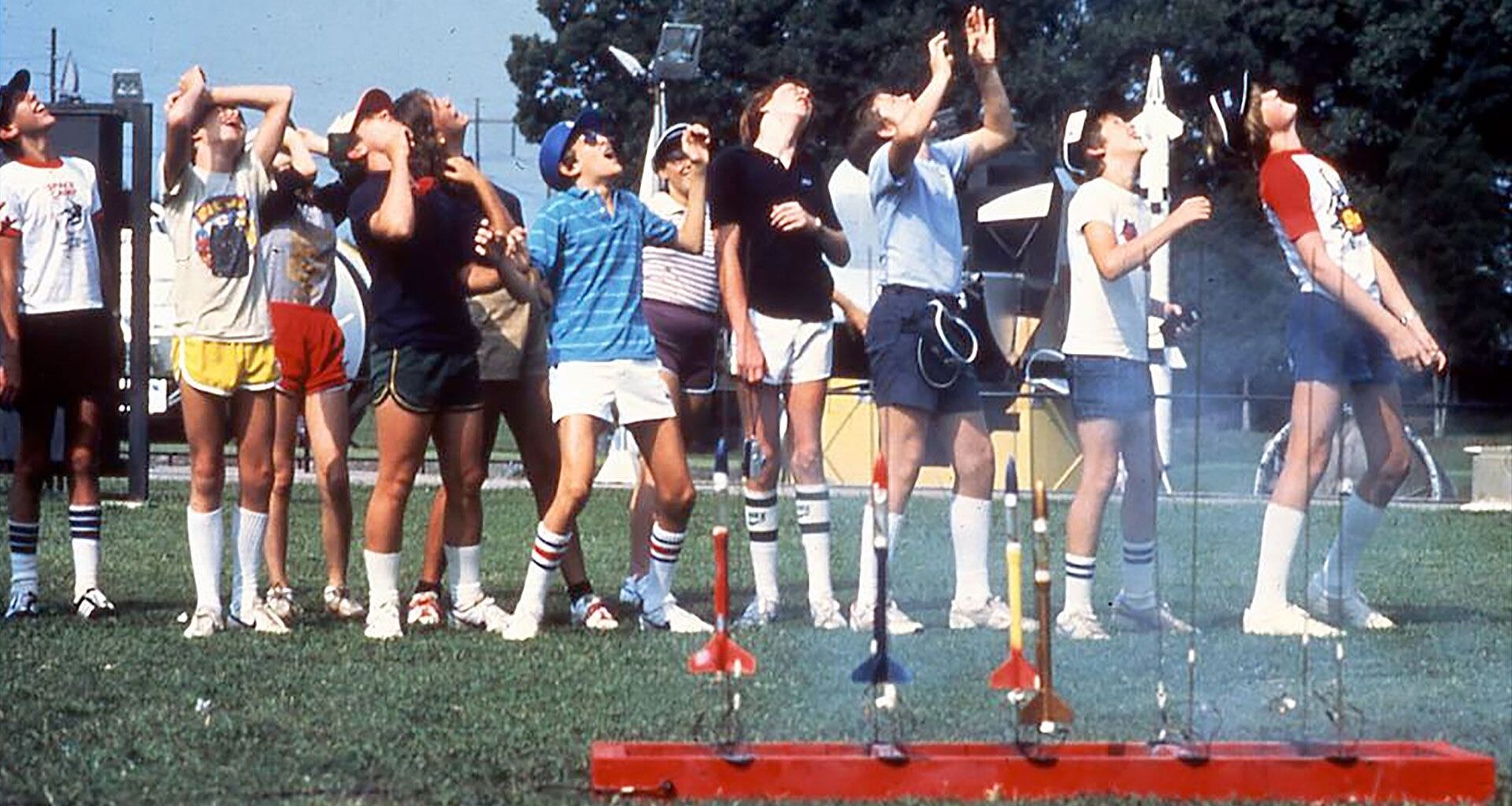 kids in multi-color t-shirts and shorts watch model rockets fly as other rockets stand ready to fly from a launch pad