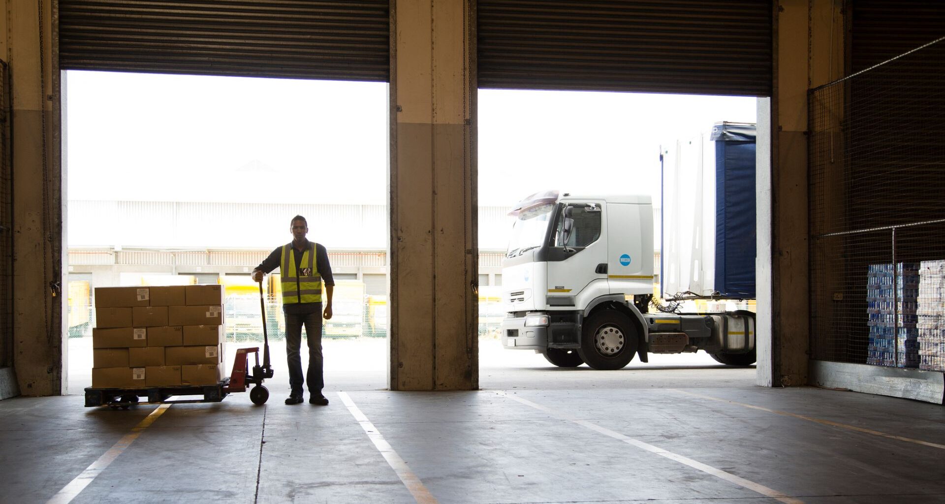A worker wearing a yellow vest stands looking into an empty warehouse with a cart full of food boxes next to him and a truck in the background