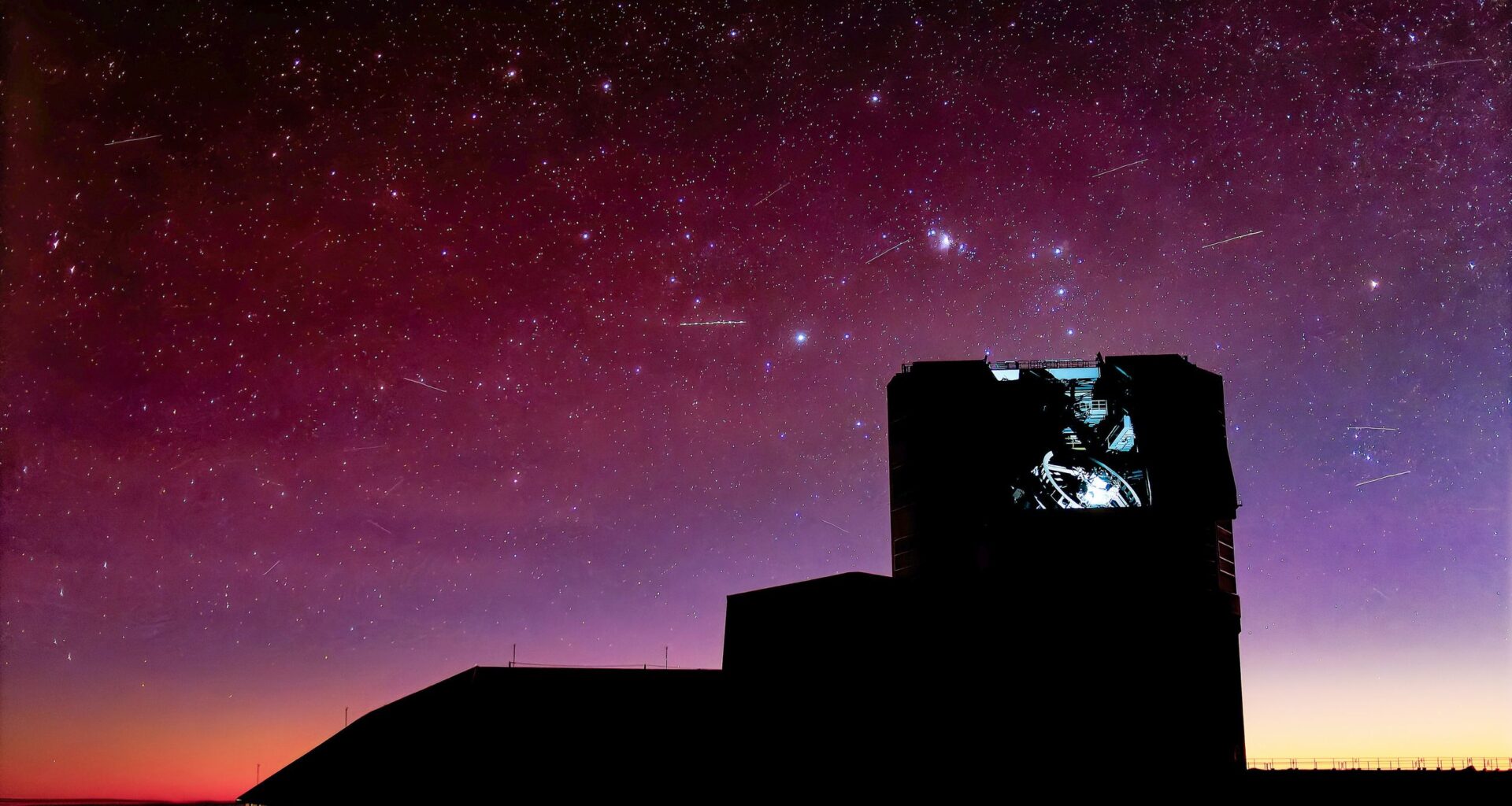 The silhouette of an observatory under a pinkish sky with lots of stars and a few streaks.