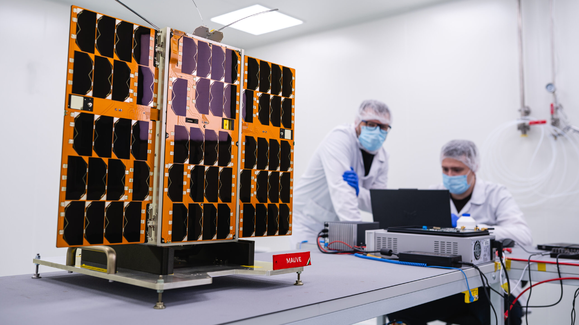 two people in cleansuits look at a rectangular shoe-box-sized satellite covered in gold foil sitting on a laboratory workbench