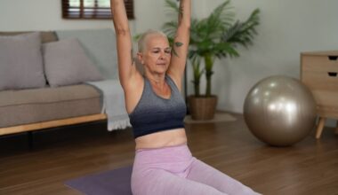 woman facing the camera sat on an exercise mat on wooden floor with her legs in front and arms stretched overhead. she's wearing light pink leggings and grey crop top. there's a sofa a golden balance ball behind her.