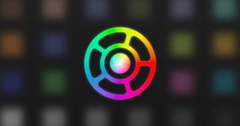 A circular logo with a black center and radiating segments, outlined in a gradient of bright rainbow colors, set against a blurred dark background featuring faint colored squares.