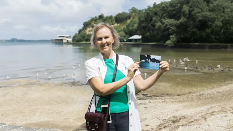 Krystene Vickers holds a historic photograph of the yacht club house at Blockhouse Bay Beach. Photo: RNZ / Yiting Lin
