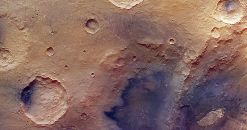 A high-resolution image of the Martian surface shows several impact craters of varying sizes on a dusty, reddish-brown landscape with some shadowed and rough textured areas.