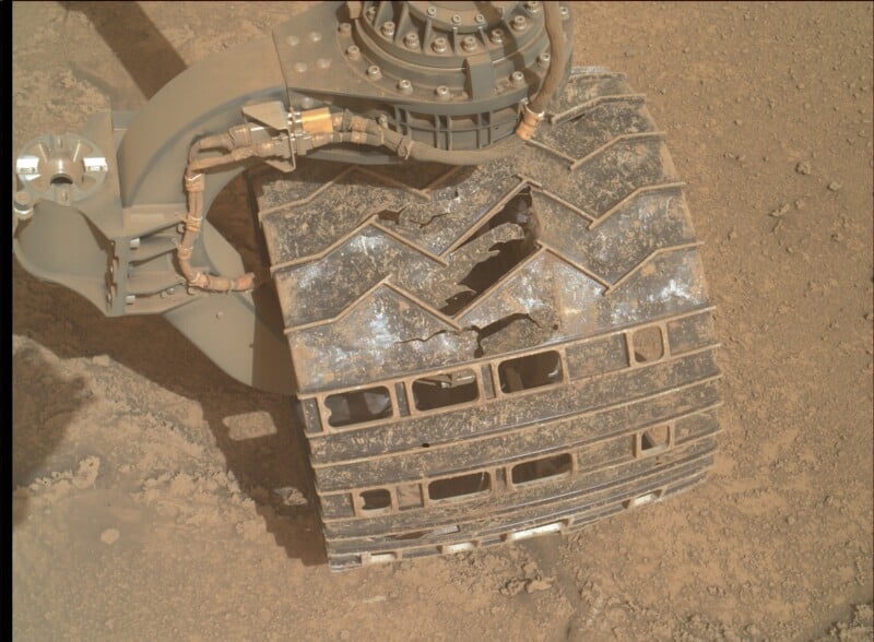 Close-up of a Mars rover wheel with multiple cracks and holes in its metal surface, resting on dusty Martian soil. Parts of the rover’s suspension and attached cables are also visible.