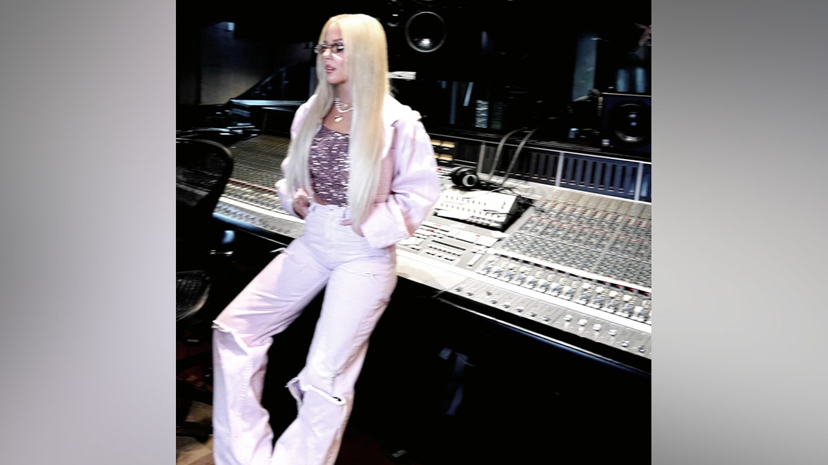 Megan Moroney with long blonde hair and glasses, wearing a pink jacket and sparkly top, leaning against a large professional soundboard in a recording studio.