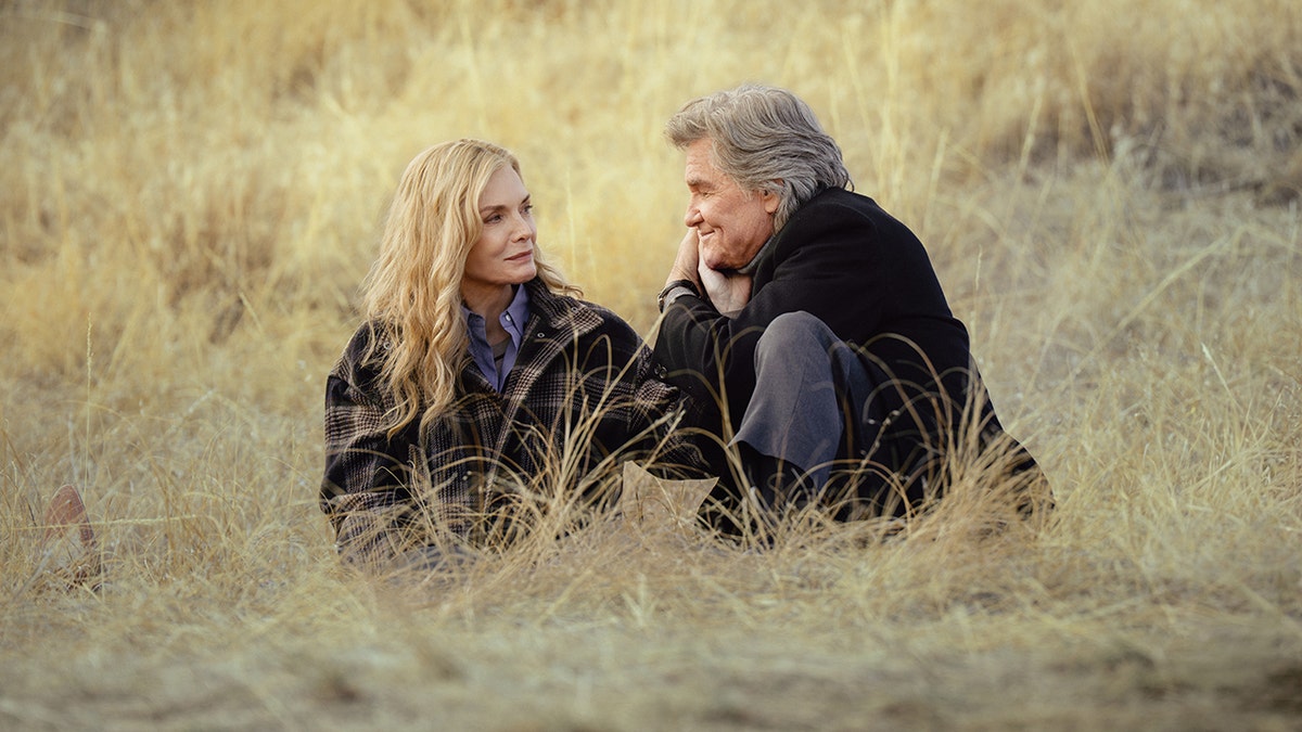 Michelle Pfeiffer and Kurt Russell sitting together in tall dry grass facing each other during a quiet outdoor moment.