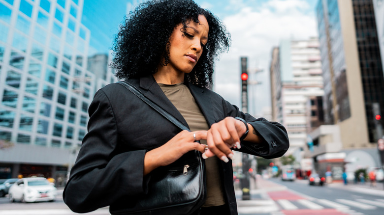 Woman dressed in business casual clothes with a purse looks down at her wrist watch while standing in the street