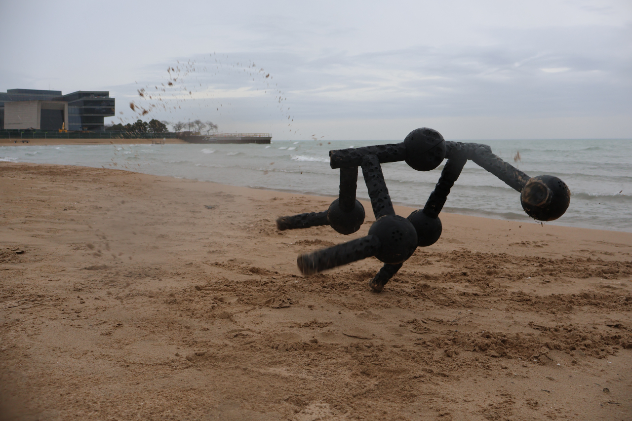 A ball-and-socket modular robot moves across a dark sandy beach with gray water behind it