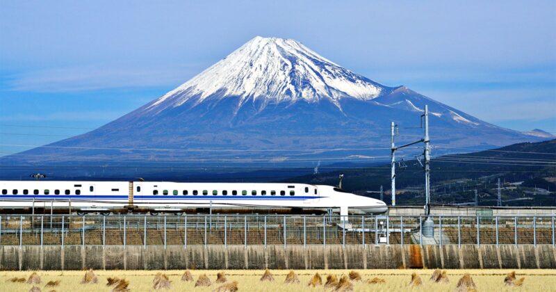 A white bullet train speeds along tracks in the foreground, with Japan’s snow-capped Mount Fuji towering in the background under a clear blue sky.
