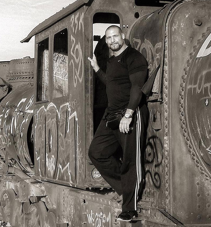Security guard posing on a graffiti-covered vintage train, wearing athletic clothing and a watch, outdoors in daylight.
