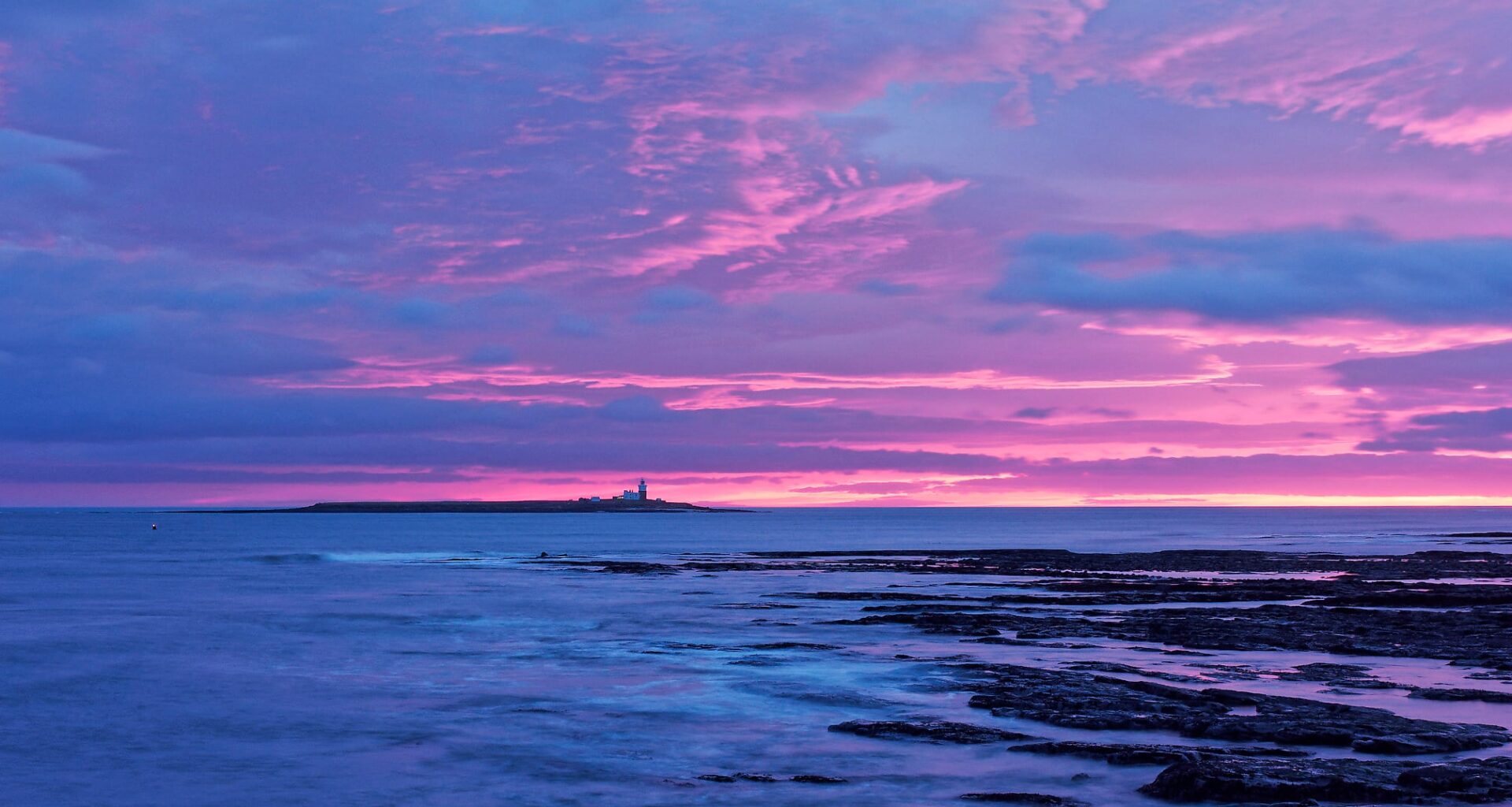 A rocky shoreline at dusk with dramatic pink and purple clouds in the sky. A small island with a lighthouse sits on the horizon, surrounded by calm ocean water reflecting the colorful sunset.