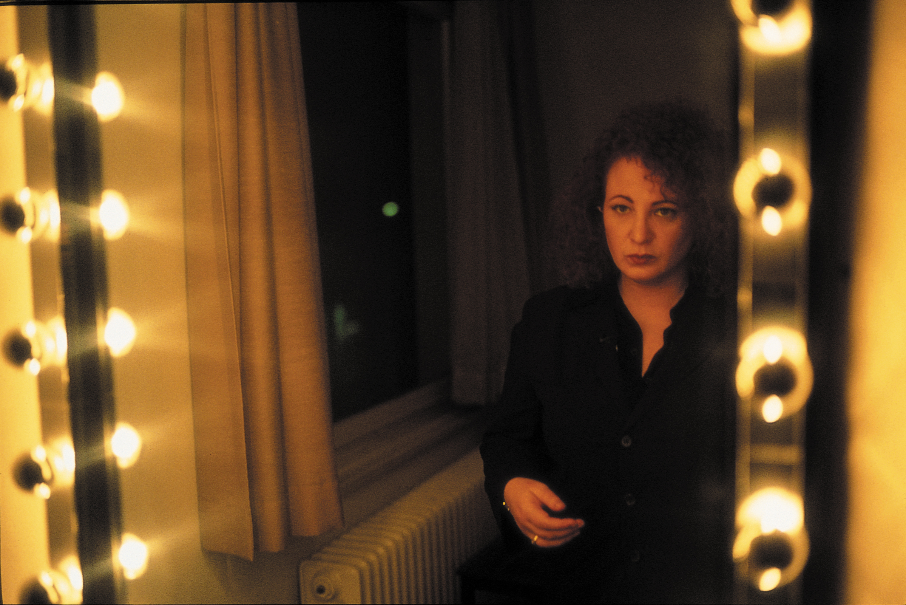 A color photograph of a woman with curly hair looking into a lighted vanity mirror in a dark room.