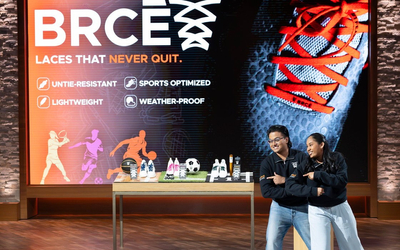 Two individuals stand on a stage in matching black tops and light pants/jeans, striking a crossed‑arms pose. Behind them is a large screen with branding that reads “BRCE” and the tagline “Laces That Never Quit.” Icons indicate product features such as untie-resistant, lightweight, sports optimized, and weather‑proof. A display table showcasing shoelaces, shoes, and sports items is placed in front of the screen.