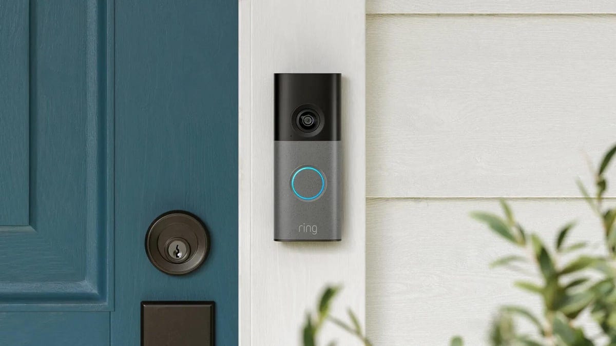 A gray Ring doorbell on white siding by a teal door.