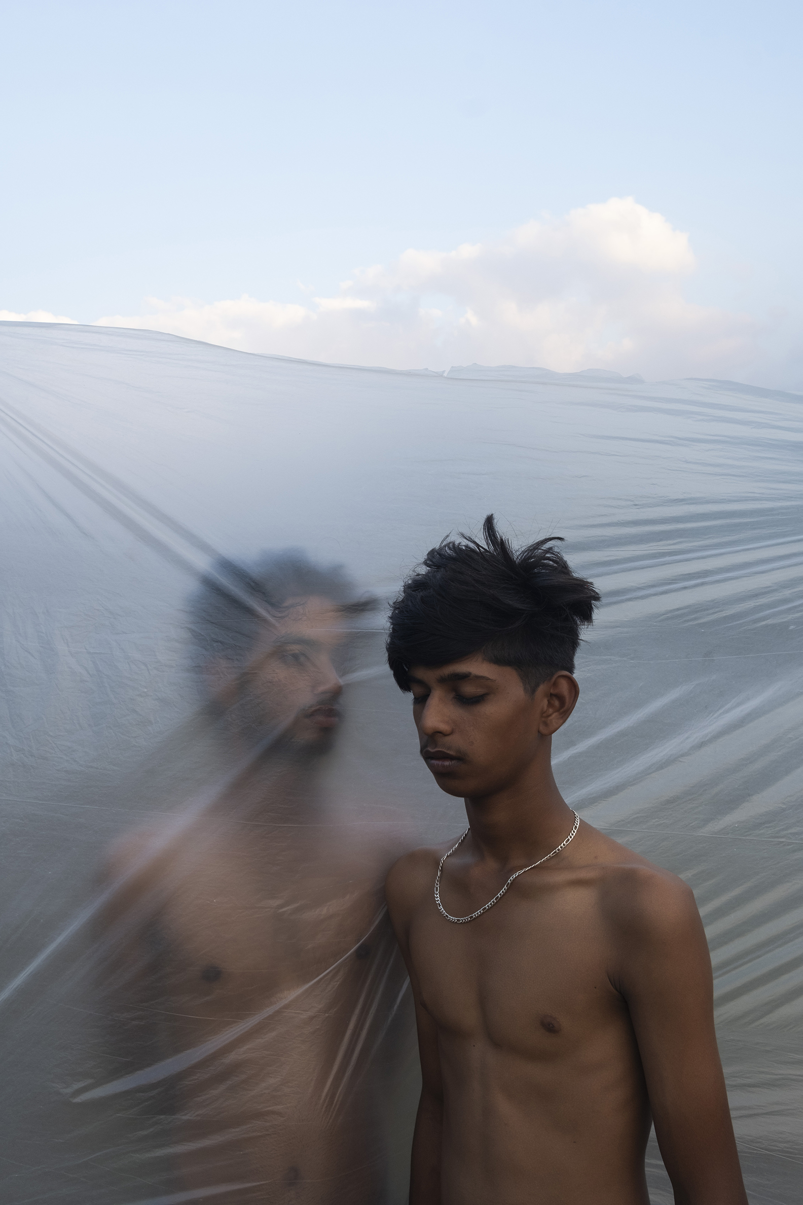 Young man with eyes closed, wearing a chain, stands against clear plastic under a blue sky. His reflection is blurred, creating a dreamy, introspective tone