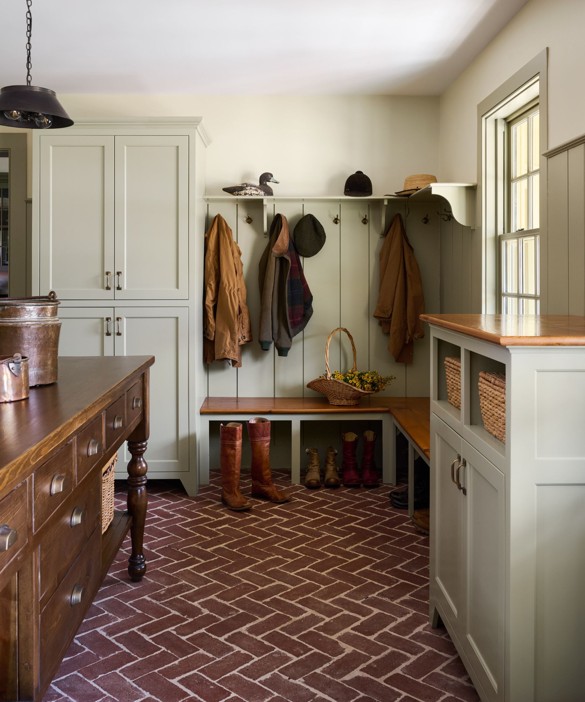 Mudroom with sage green painted cabinetry and panelling on walls, terracotta brick tiled floor, mahogany table with storage, and a built-in bench with boots underneath