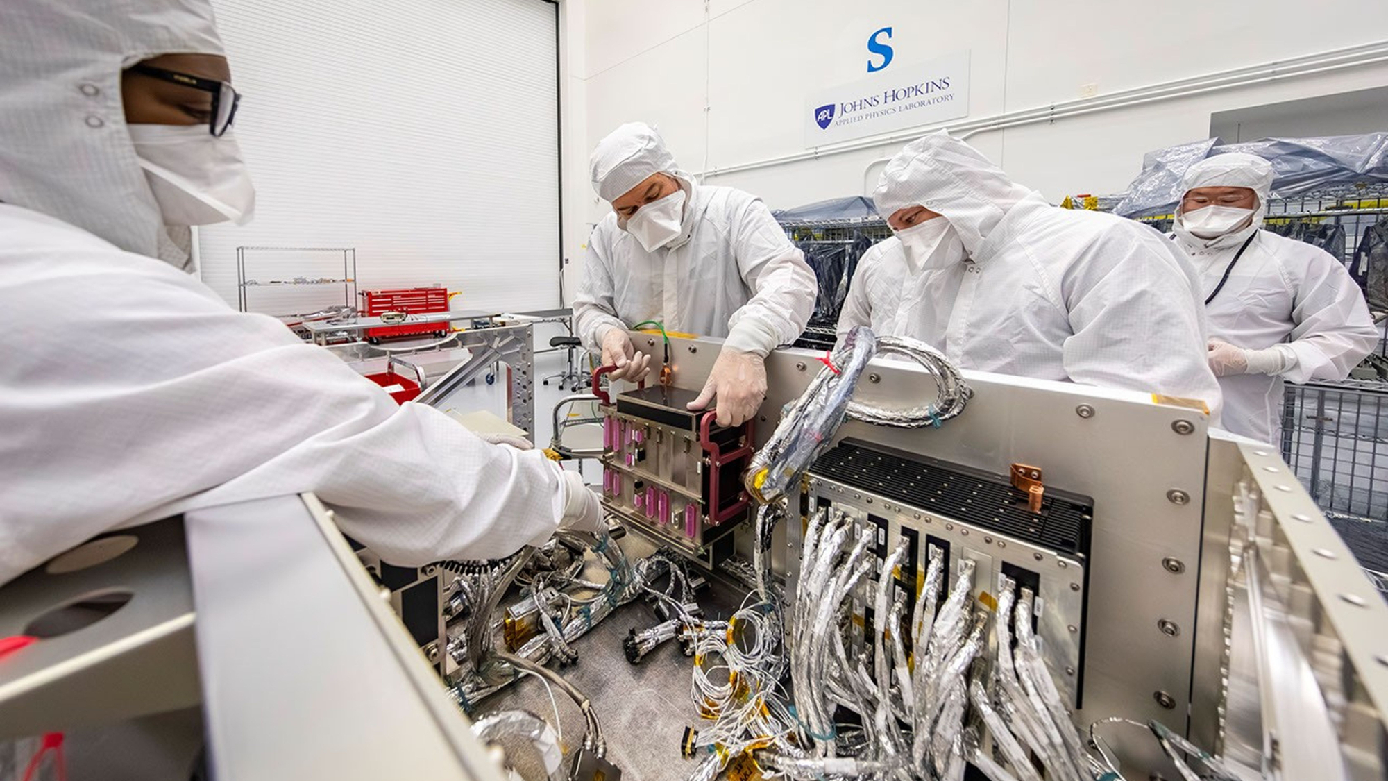 four people in white protective clothing work on a complex piece of electrical equipment inside a white-walled scientific lab