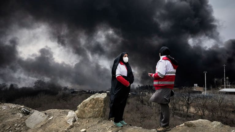 Two women from the Iranian Red Crescent Society stand as a thick plume of smoke from a US-Israeli strike on an oil storage facility late Saturday rises in the sky in Tehran, Iran, Sunday, March 8, 2026.