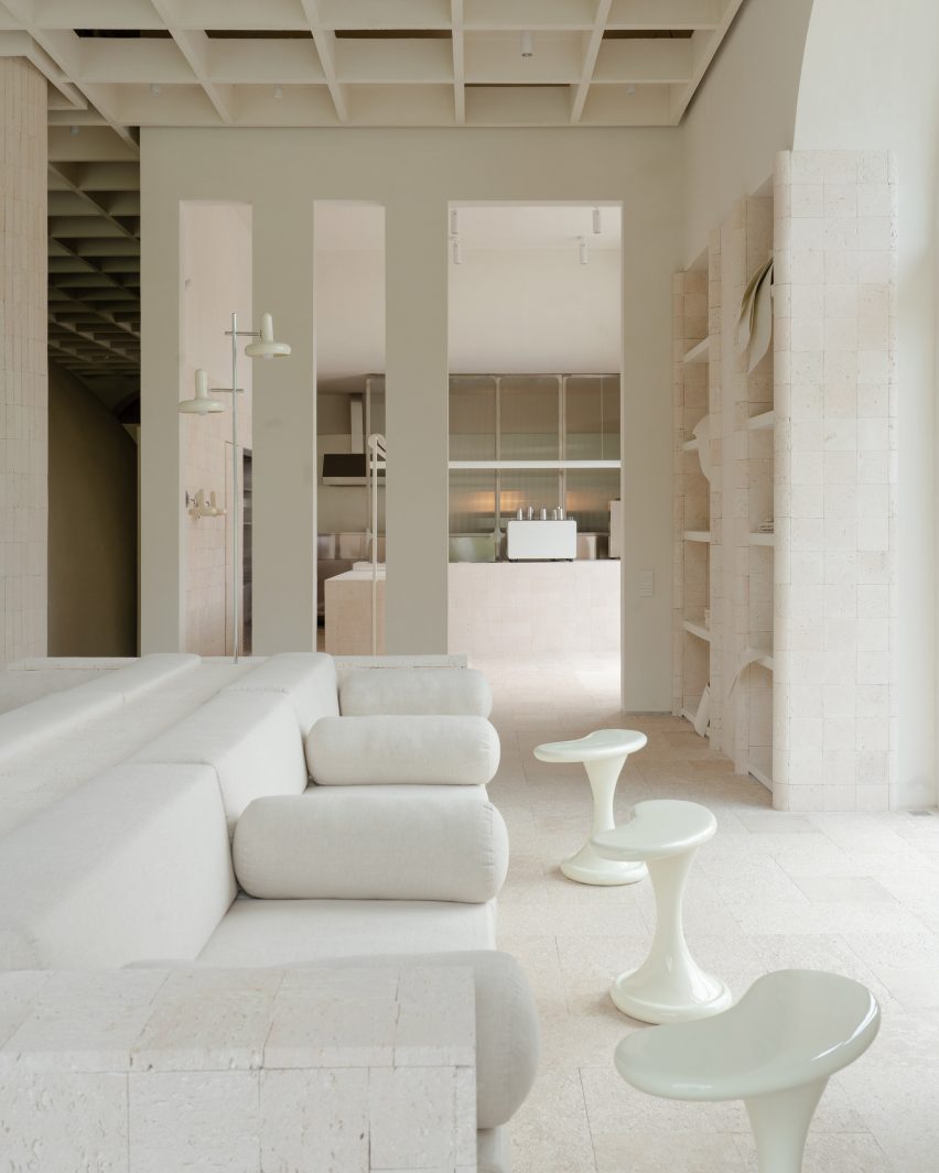 Image of a neutral, off-white interior with a low boxy sofa and three free-form side tables. Large rectangular archways in the background showcase a cafe counter and service area.
