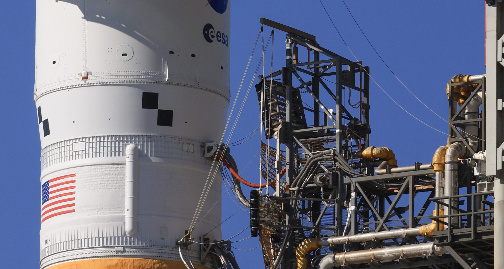 A section of a rocket seen with multiple lines, or umbilicals, connecting it to eqiupment on a tower.