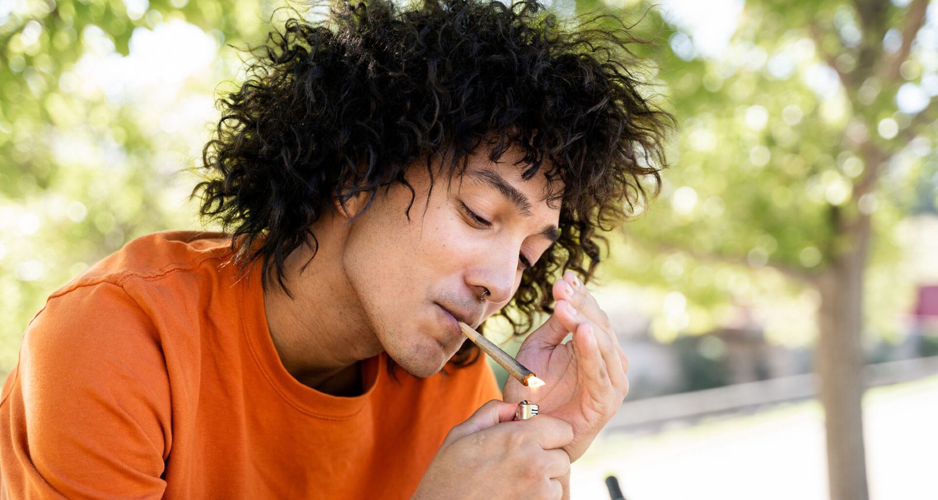 A man with long wavy black hair wearing an orange shirt lights a joint with a lighter