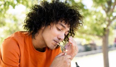 A man with long wavy black hair wearing an orange shirt lights a joint with a lighter